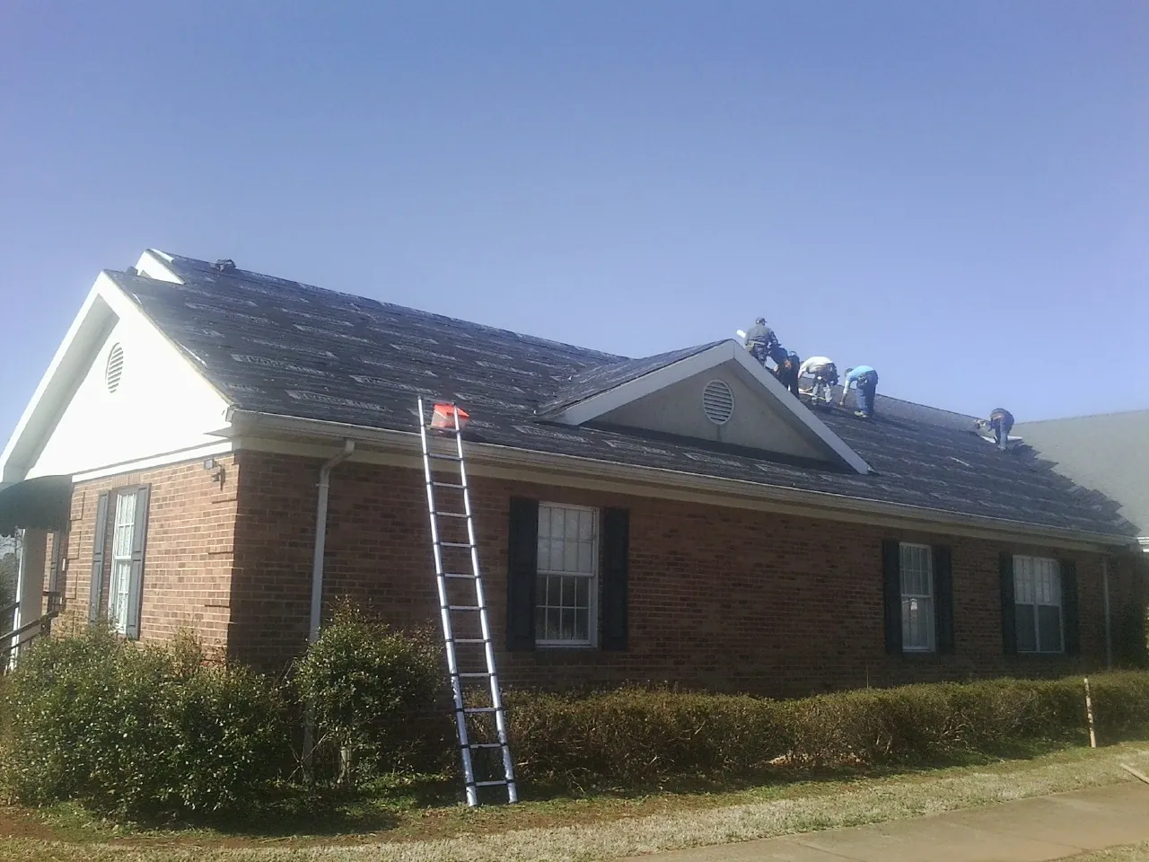 Crew tearing shingles off a brick ranch roof with a ladder set — Metal Roof Repair work by our crew in Orfordville