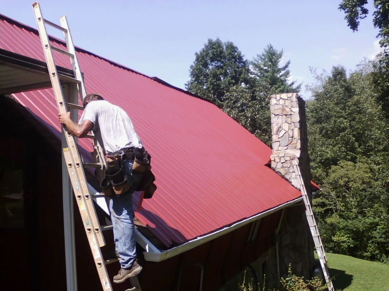 Roofer on a ladder beside a red metal roof and stone chimney — Soffit & Fascia Repair by Roofing Brothers in Orfordville