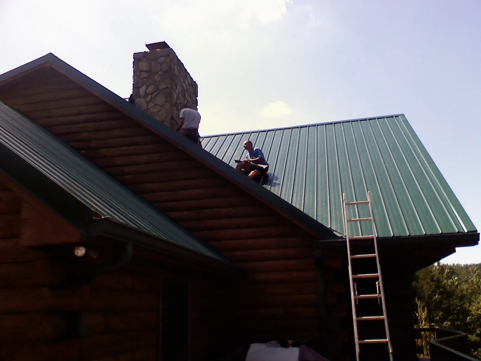 Roofers installing green metal panels beside a stone chimney on a log home in Orfordville